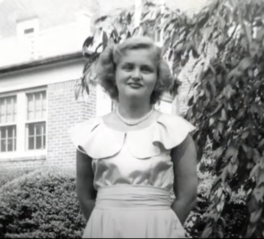 A young Mildred Oberkotter stands in front of a brick structure and green foliage, smiling with her hands behind her back.
