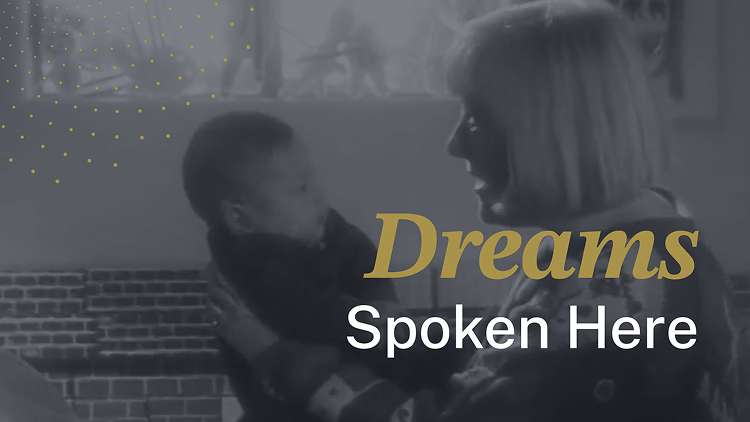 A woman cradles a baby in her arms and speaks to it. The baby, who is wearing a hearing device, looks back at the woman. The text “Dreams Spoken Here” appears on the image.  