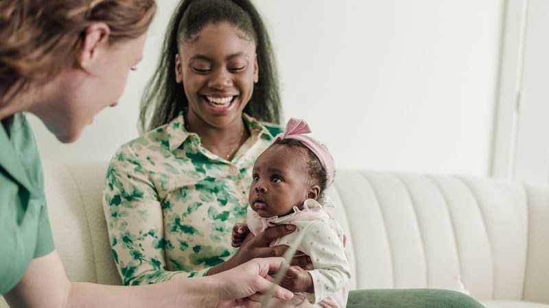 Picture of a mom smiling and holding her baby on her lap while sitting on a couch and a nurse is speaking to them both.
